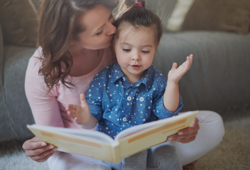 A mother sits on the floor of her living room with her young daughter in her lap and reads a children's book.